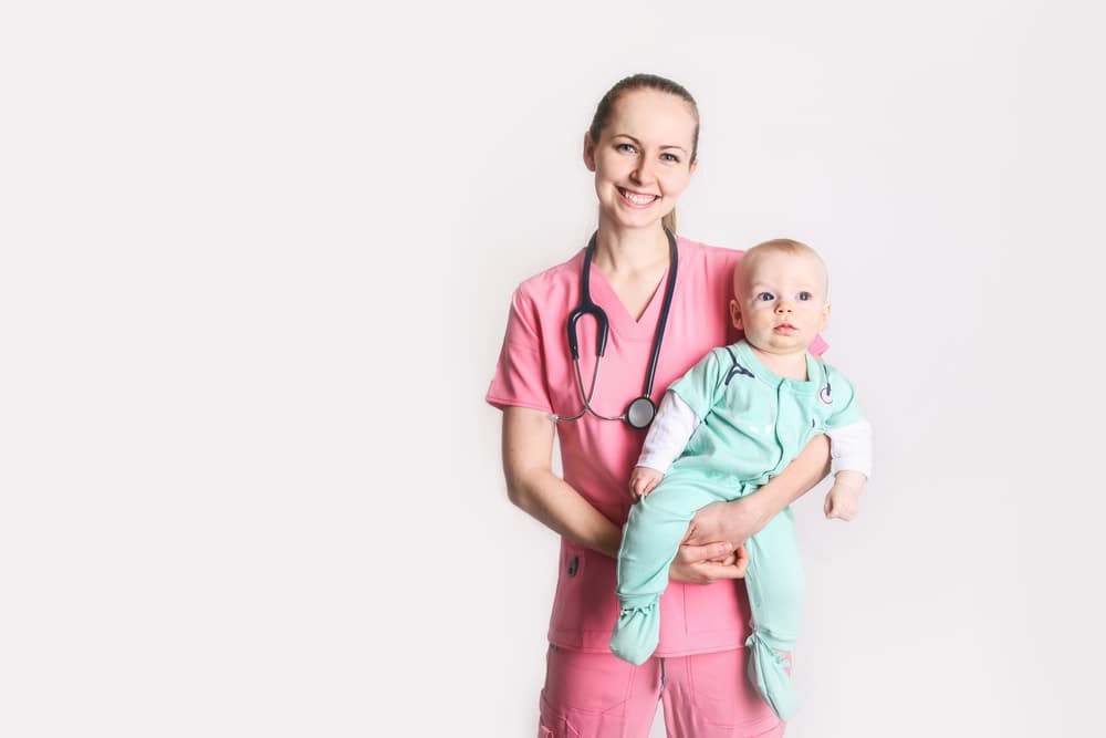 A smiling travel nurse, wearing pink scrubs and a stethoscope, hold her child who is wearing a onesie with a medical print design. The nurses hair is pulled back in a ponytail and she appears to be happy and content.