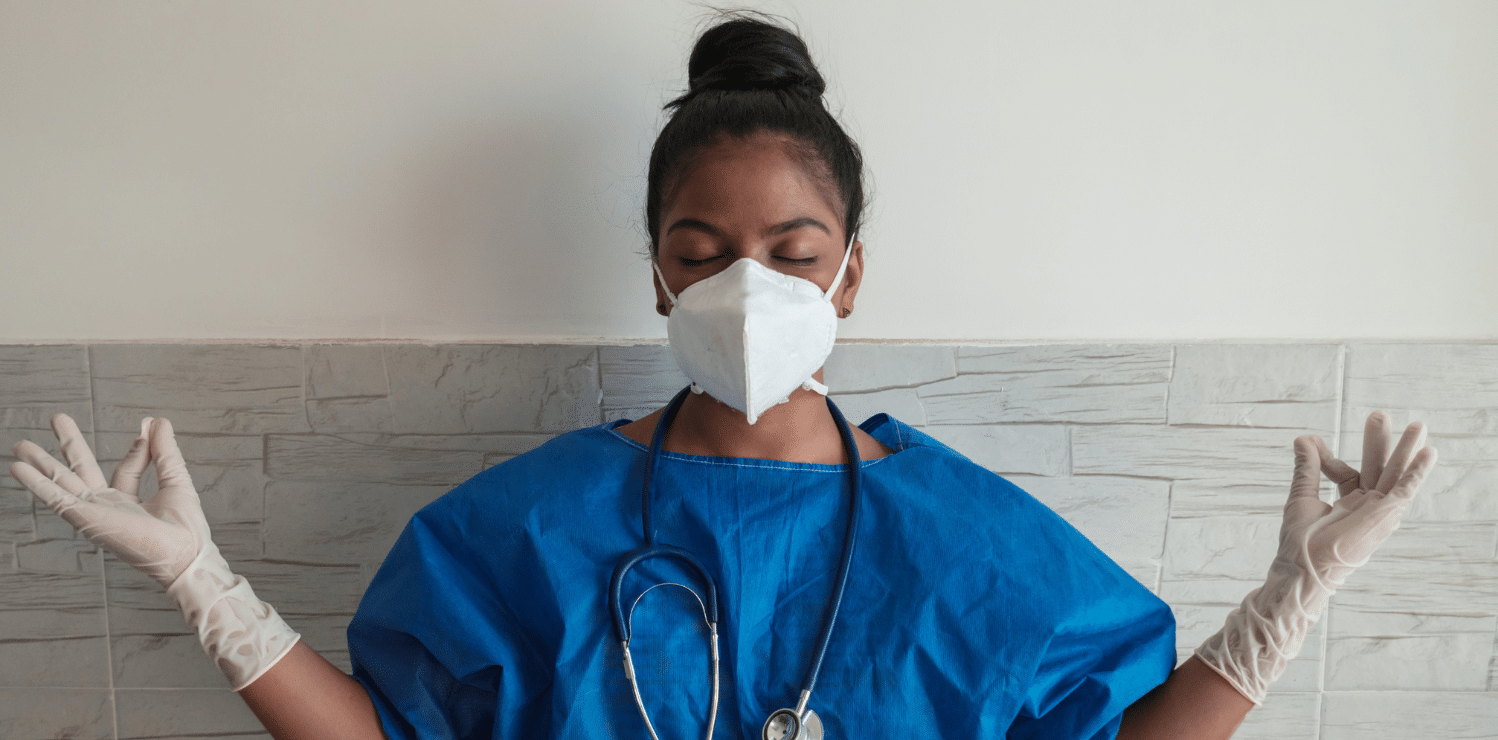 Nurse wearing blue scrubs, a face mask, and gloves. She is meditating with her eyes closed, showing that she is taking care of her mental health while on the job. The background is a clean and calming medical facility, which suggests that she is taking a break during a busy workday to practice mindfulness.