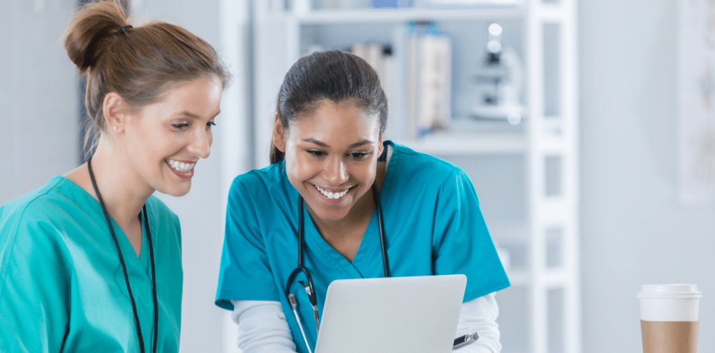 Two female travel nurse friends are smiling while looking at a laptop together in a medical setting. They are both wearing scrubs and appear to be enjoying their assignment.