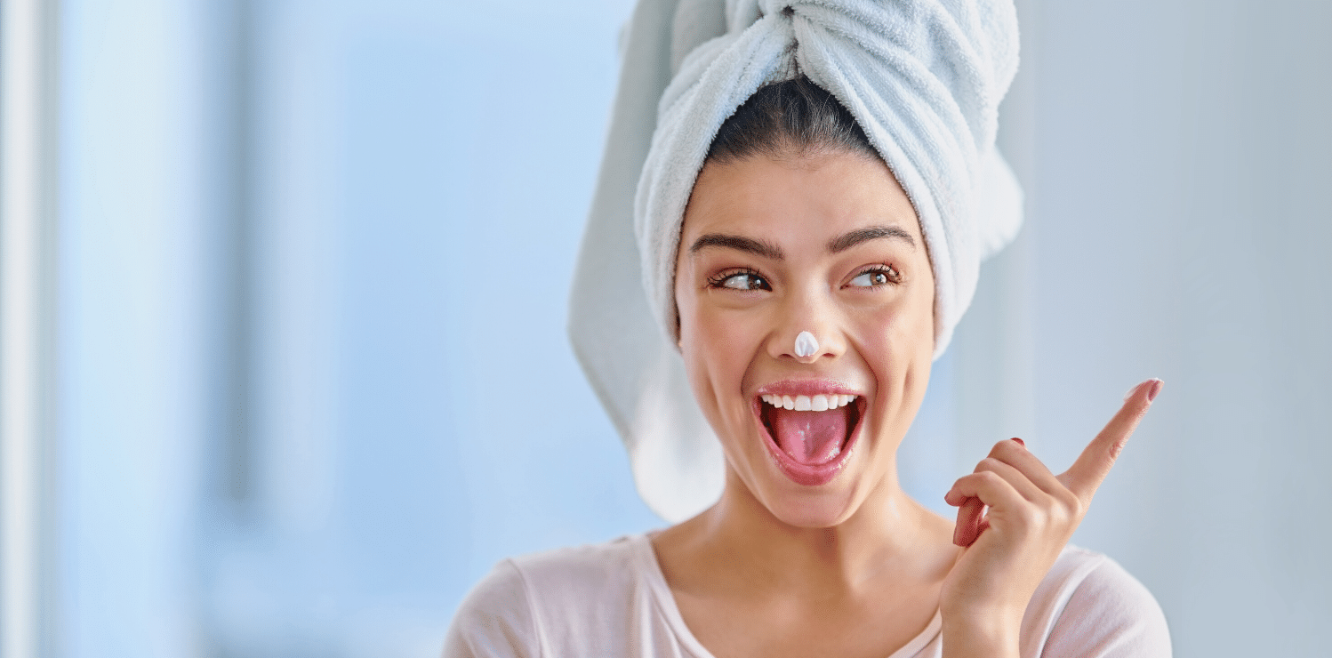 A woman with her hair wrapped in a towel smiles while applying skincare. She is wearing a skincare treatment mask on her nose.