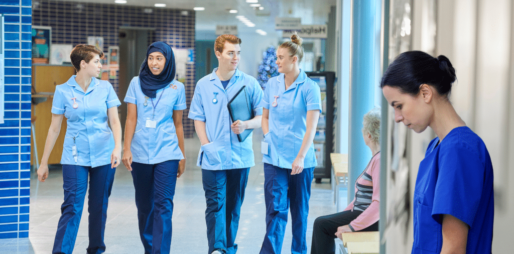 A travel nurse experiencing loneliness on her assignment stands alone in a hallway, looking isolated and depressed, while in the background a group of other nurses walk together in a medical setting, engaged in conversation.