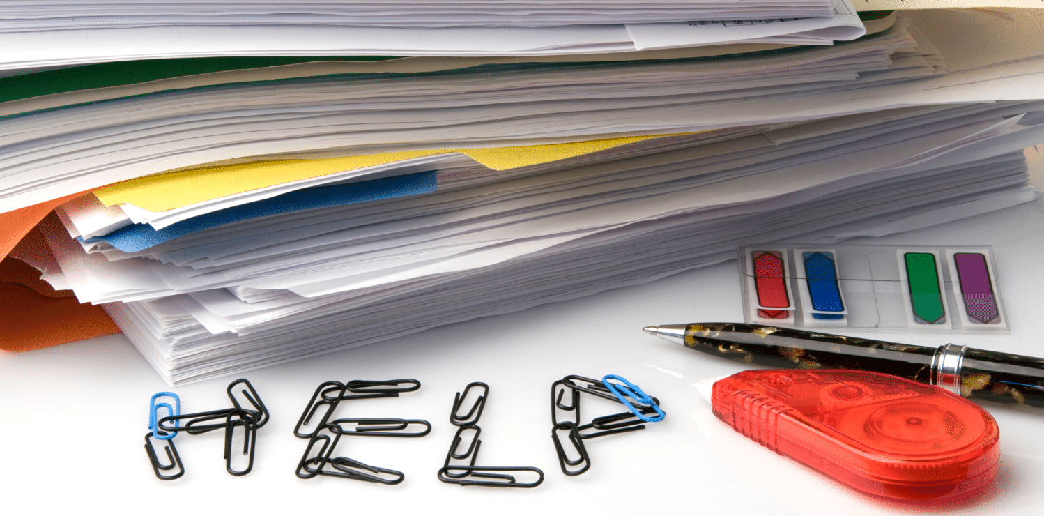 A stack of credentialing and onboarding paperwork on a desk with a pen, white out, sticky tabs and paper clips arranged to spell out the word "help".