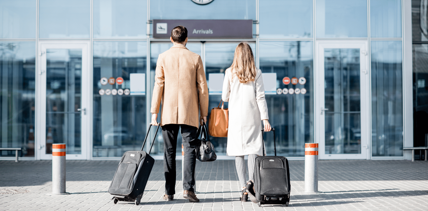 Spouses walking into the airport with their backs turned to the camera, each pulling their luggage behind them. Both are dresses professionally, suggesting that they may be traveling for work or a professional engagement.