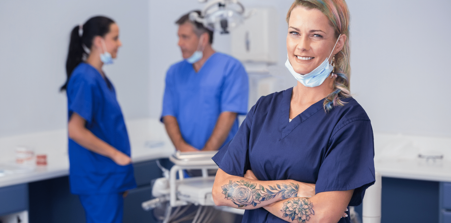 Medical professional wearing blue scrubs, with a face mask draped under her chin, showing her colorful pastel rainbow hair, floral tattoos, and piercings. She has a warm smile on her face and her arms crossed, confidently displaying her unique style. In the foreground, two nurses are talking while working, highlighting the collaborative and supportive nature of the healthcare industry.