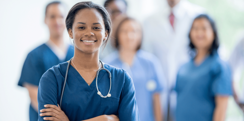 Confident travel nurse standing out from a group of other nurses. She is wearing blue scrubs and a stethoscope around her neck, with her arms crossed and a smile on her face.