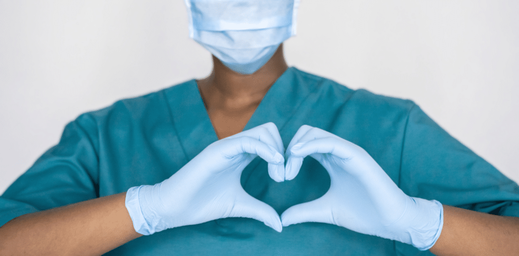 Nurse in blue scrubs and face mask expressing joy and optimism, forming a heart symbol with her hands over her chest, celebrating the WHO's announcement of the end of COVID-19.