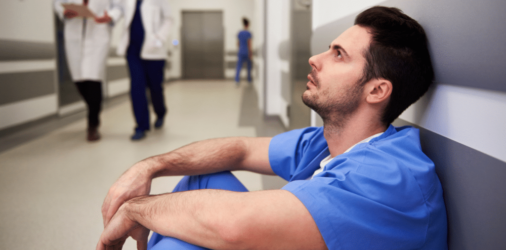 Male nurse in blue scrubs sits with his back against the wall of a medical facility hallway, appearing stressed. In the background, other healthcare professionals can be seen walking by.