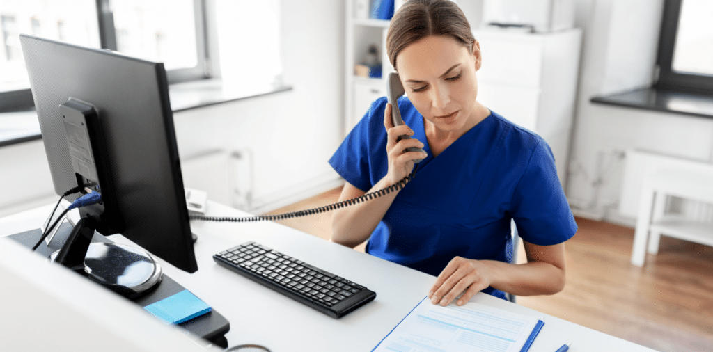 Travel nurse wearing blue scrubs sitting at her desk, reviewing paperwork while on the phone.