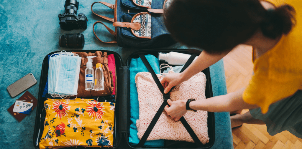 A woman is packing a large suitcase for her travel nurse assignment. The suitcase is open and filled with neatly folded clothes, medical supplies, and personal items. The woman is wearing a bright yellow top and sage green skirt, indicating her professional role. She carefully arranges items in the suitcase, making efficient use of space. Surrounding her are travel essentials such as face masks, hand sanitizer, a passport, and her cellphone. The room is well-lit with natural sunlight streaming through a window, creating a warm and inviting atmosphere. The woman's confident and focused demeanor suggests her readiness to embark on her new nursing adventure.