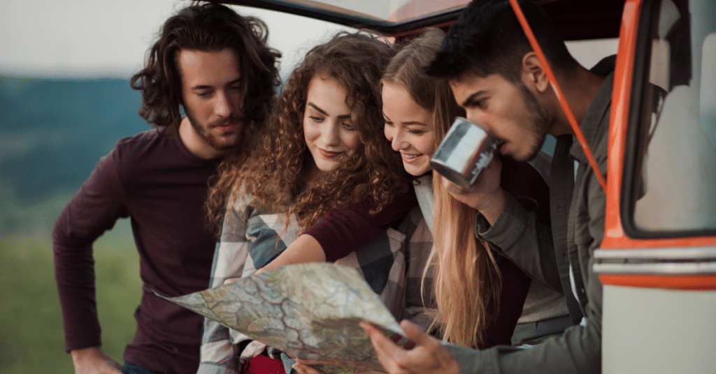 Vibrant young friends embracing adventure in a retro minivan during American Adventures Month. They joyfully navigate a road trip through the countryside, engrossed in studying a map together, making memories to cherish for a lifetime.