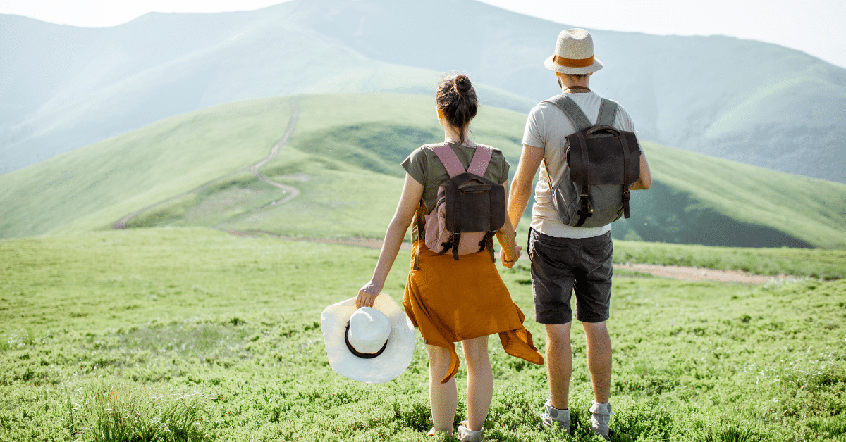Young travel nursing couple savoring a scenic moment together, backpacks on, immersed in the beauty of green mountains, captured from behind. This image captures the true essence of travel nursing as a couple.