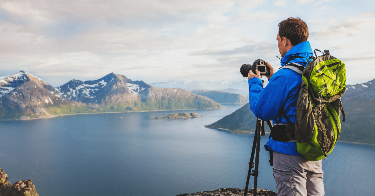 Travel nurse setting up a tripod to capture a scenic photograph outdoors, with a breathtaking view of a serene lake and majestic mountains in the background. Learn the art of capturing stunning moments during your travel nursing assignments.