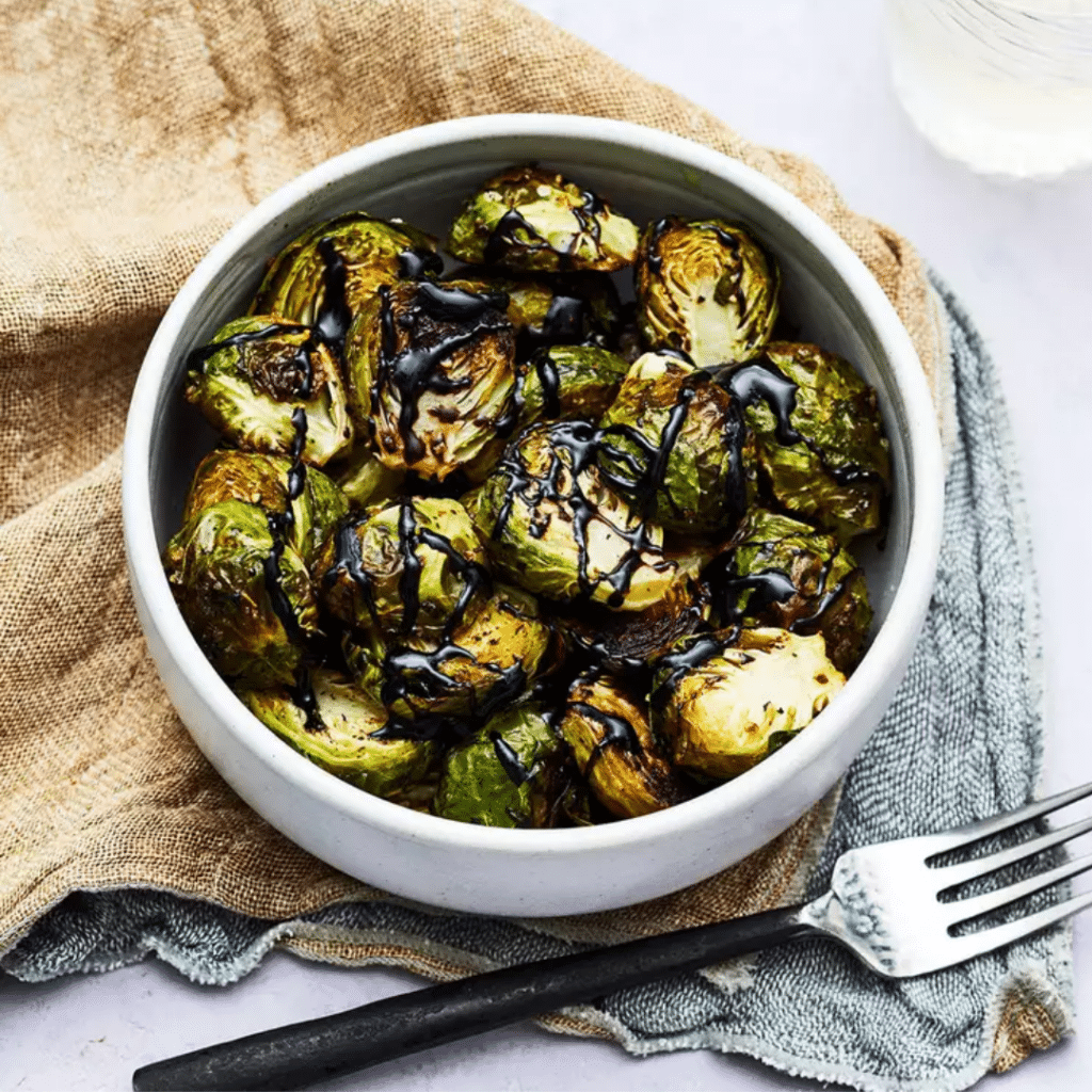 A bowl of Honey-Balsamic Brussel Sprouts, beautifully roasted to perfection, sits on a table. The dish is garnished with a drizzle of honey and balsamic glaze, creating a visually appealing and delicious Thanksgiving meal. This dish is an ideal choice for travel nurses working on temporary assignments, offering a taste of comfort and festivity during their busy schedules.