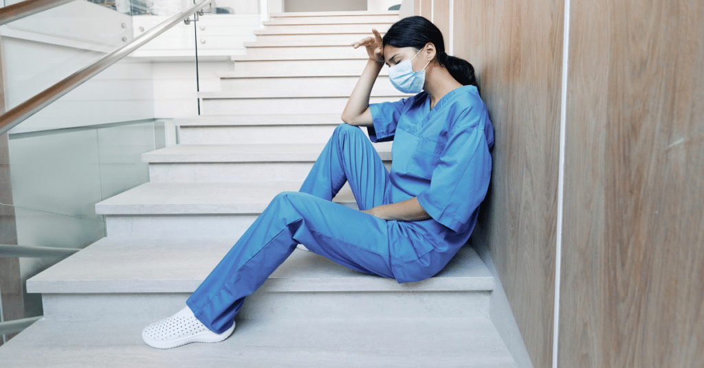 A nurse, dressed in blue medical scrubs and wearing a face mask, is sitting at the bottom of a staircase. She appears tired or stressed, with one hand resting on her head, conveying a sense of feeling off during her nursing shift. The setting suggests a brief moment of pause in a demanding work environment.