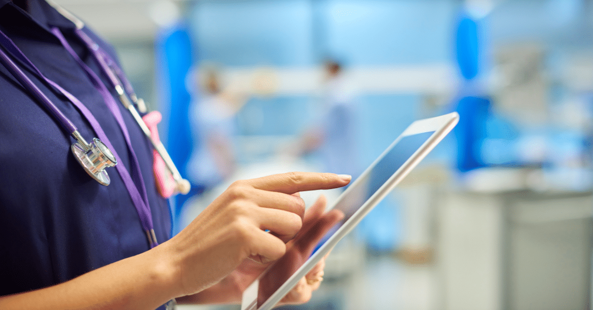 A waist-up view of a nurse in a busy hospital environment, focused on a tablet in her hands. The nurse's face is not visible, emphasizing the device which displays various apps suitable for travel nurses. The bustling hospital backdrop suggests a fast-paced work setting.