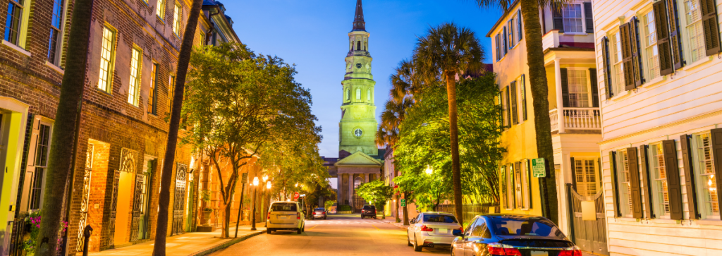 A scenic view of Charleston, South Carolina, focusing on the historic French Quarter at twilight. The landscape showcases the unique blend of historic architecture and lush greenery typical of this area, with the soft hues of twilight casting a serene glow over the buildings. The streets are quiet, reflecting the area's underrated charm, making it an appealing destination for travel nurses seeking a picturesque and peaceful environment.