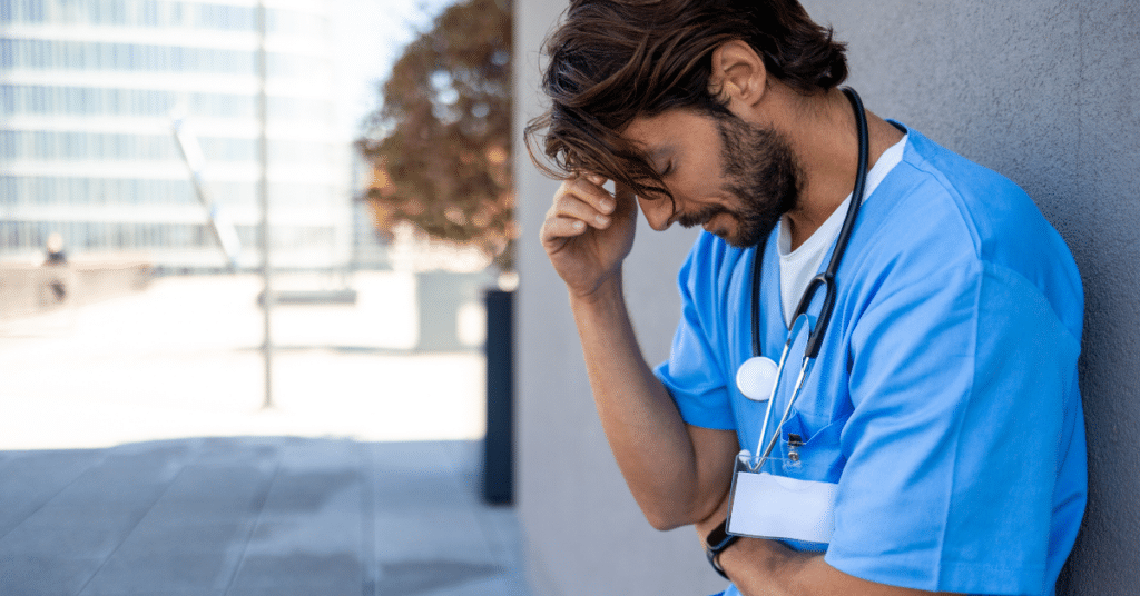 Image of a stressed male nurse taking a break outside of a hospital. He is standing with his hand on his brow, looking downwards, reflecting a sense of exhaustion or concern.