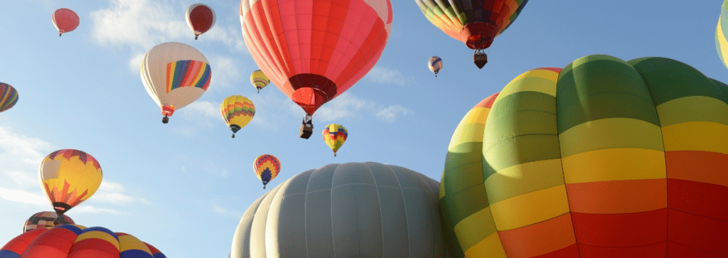 This image features a vibrant scene from the ballooning festival in Albuquerque, New Mexico, near Santa Fe. It showcases a collection of colorful hot air balloons of various shapes and sizes, floating gracefully against a clear blue sky. The festival atmosphere is lively and picturesque, making it an appealing and underrated destination for travel nurses looking for unique experiences.