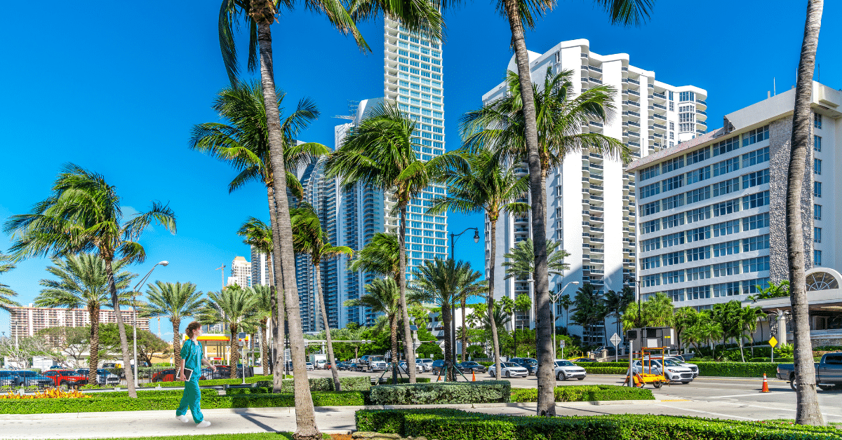 Street view of Sunny Isles in Miami, Florida, featuring a travel nurse in blue medical scrubs standing with a tablet. She appears content and pleased with her job as a travel nurse in Florida.
