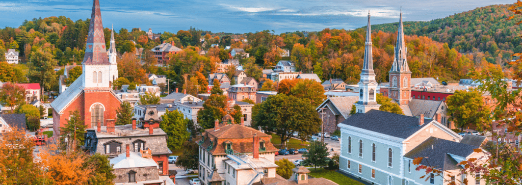 The skyline of Montpelier, Vermont, USA, captured during twilight. The city's buildings are silhouetted against a sky that blends the soft hues of sunset. Street lights and windows in the buildings begin to glow, adding a twinkling effect to the urban landscape. In the background, the rolling hills and trees characteristic of Vermont can be seen, creating a contrast between nature and the cityscape. The overall atmosphere is serene and picturesque, typical of a small American town at dusk. Vermont is an underrated destination for travel nurses.