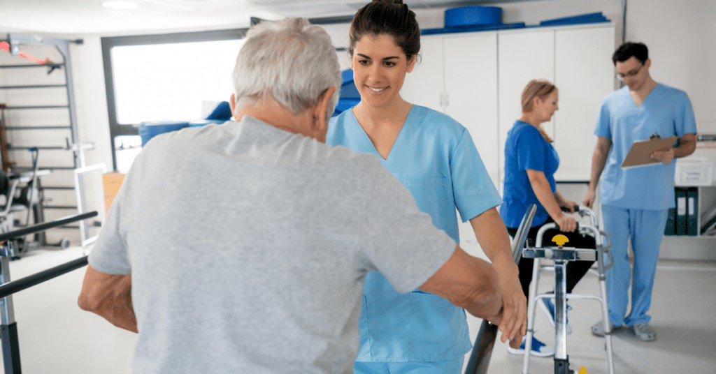 Patients at an occupational therapy facility are each working with a therapist, using parallel bars and aid walkers as part of their rehabilitation. The therapists are attentively guiding the patients, demonstrating techniques for mobility and balance. The scene reflects the busy environment of managing heavy caseloads, with each occupational therapist dedicated to providing individualized care and support to their patients.